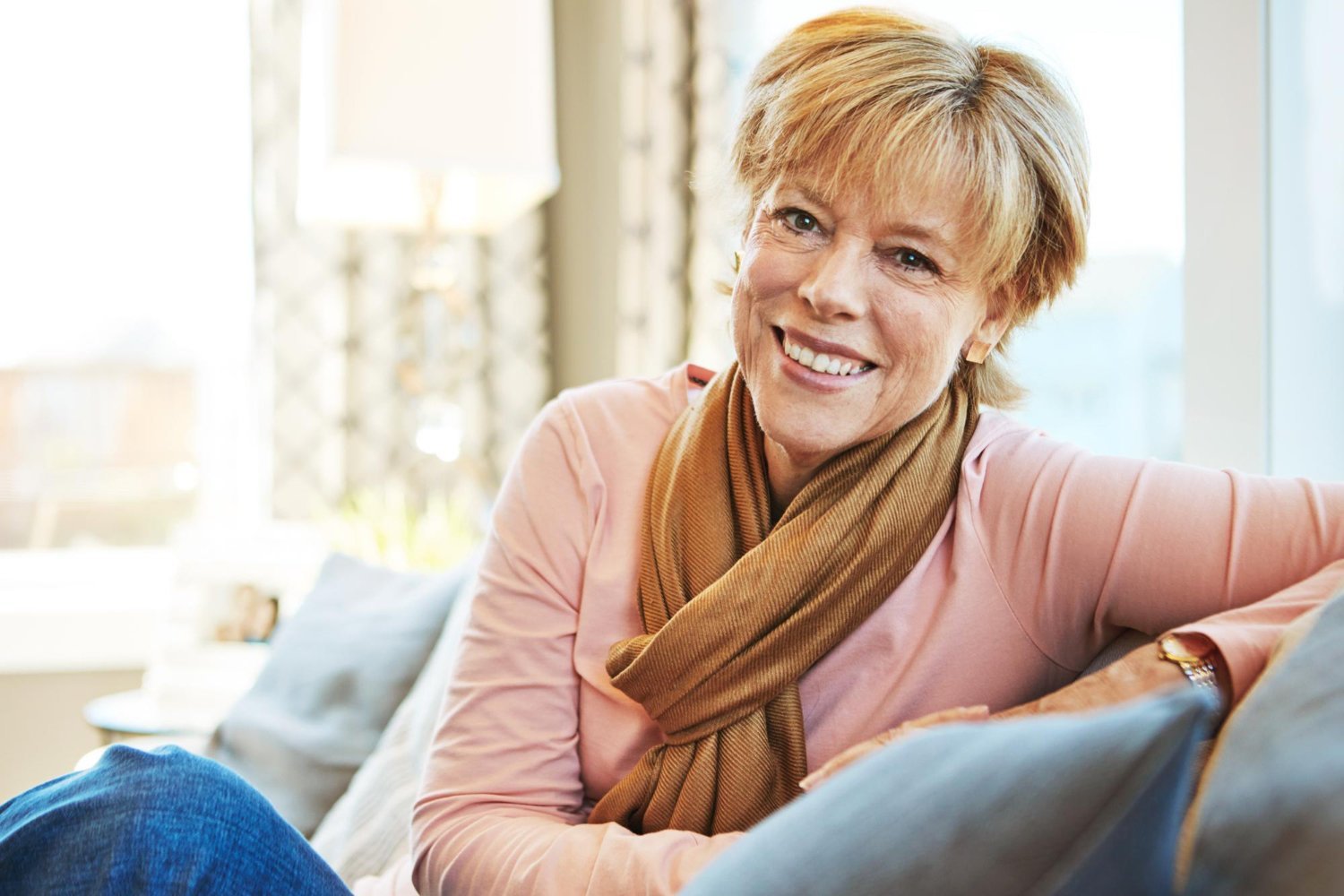 Smiling woman with short blonde hair wearing pink cardigan sitting on couch by window